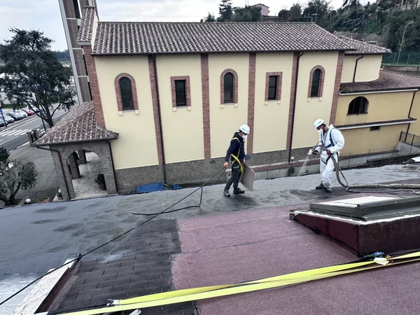 Due operatori con casco e imbracatura durante l’applicazione a spruzzo su copertura inclinata con lucernario.