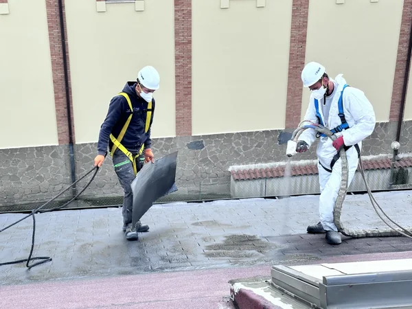 Applicazione a spruzzo sulla copertura inclinata con operatore e assistente che protegge l’area vicina.
