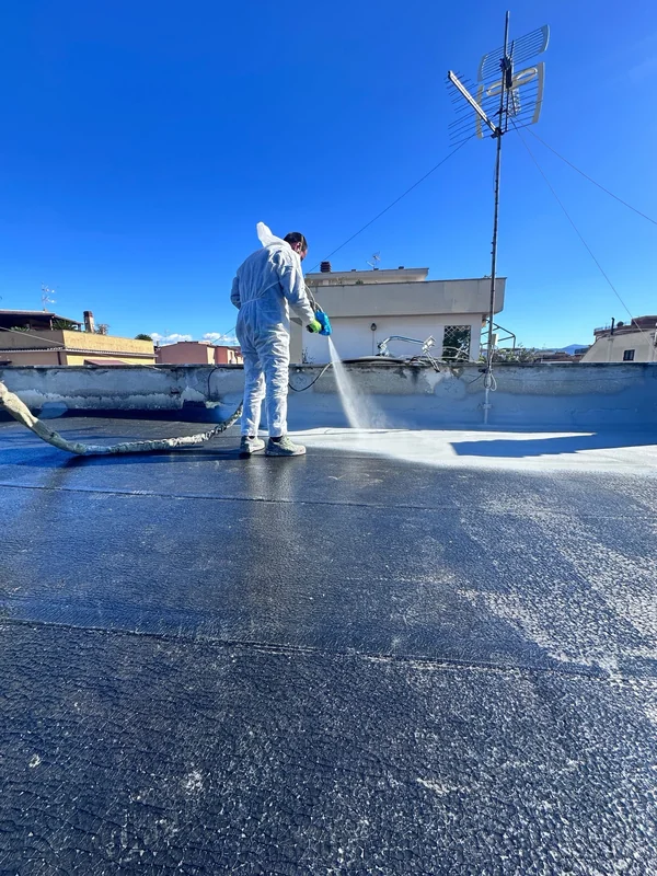 Applicazione a spruzzo sulla copertura piana, con operatore in tuta e raccordi lungo i parapetti.