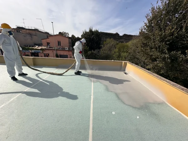 Operatore con maschera e tuta protettiva durante l’applicazione a spruzzo sul raccordo tra copertura e parapetto.